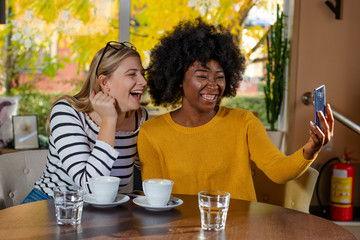 Two multi ethnic girl friends enjoying coffee together in a coffee shop, sitting at a table, chatting and laughing out loud while taking a selfie with a smartphone.