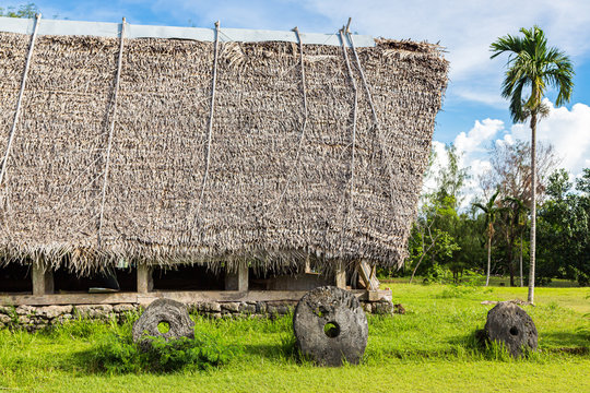 Traditional thatched yapese men's meeting house called faluw or fale and a bank of three historic megalithic stone money rai in front of it. A high coconut palm. Yap island, Micronesia, Oceania