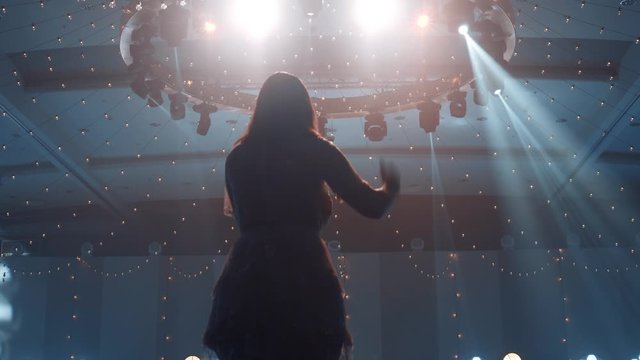 Silhouette of a young woman singer on a background of lights, spotlights, rays of light. A woman sings on stage and moves her hands to the rhythm of the song. Taken from the back. 4k footage.