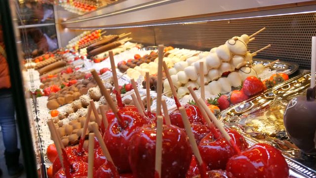 Various fruits on a wooden skewer stick in caramelized glaze made of milk black white chocolate. Apples in icing on the counter of the Christmas market. Street food