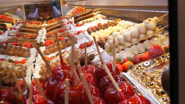 Various fruits on a wooden skewer stick in caramelized glaze made of milk black white chocolate. Apples in icing on the counter of the Christmas market. Street food