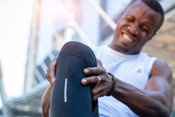 African American male runner bends over clutching his knee while in intense pain from an acute knee...