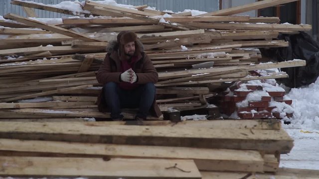 a bearded man in a hat with earflaps and a fur coat is freezing in the winter on the street against the background of boards on a construction site