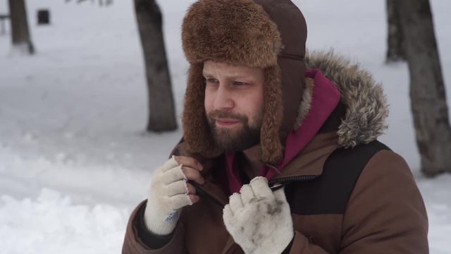 a bearded man in a hat with earflaps and a fur coat is freezing in the winter on the street