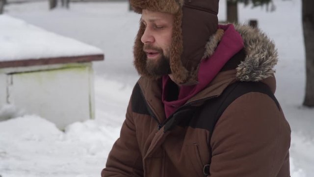 a bearded man in a hat with earflaps and a fur coat is freezing in the winter on the street