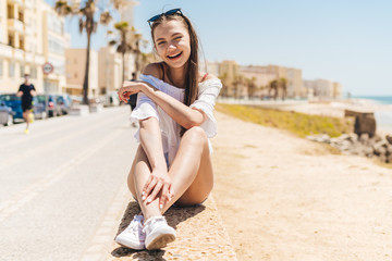 girl laughs, resting her elbow on her knees, sitting on the parapet on the background of the beach, houses and the highway