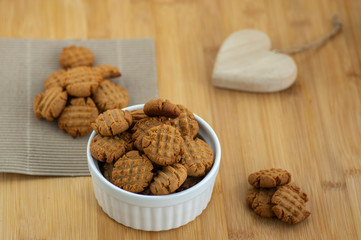 Very tasty peanut butter biscuits on bamboo wooden board in white baking bowl and decorative wood heart, golden baked healthy