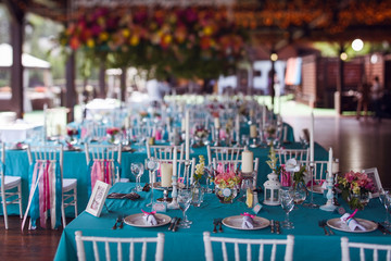 Table setting for a wedding party in pink and turquoise colors. White chairs with silk ribbons. Luxury decor. Plates, glasses, cutlery and candles. Vases with flowers.