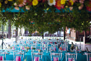 Table setting for a wedding party in pink and turquoise colors. White chairs with silk ribbons. Luxury decor. Plates, glasses, cutlery and candles. Vases with flowers.