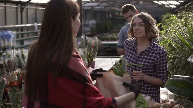 A young girl purchasing organic food in local greenhouse, recieving her grocery in paper bag and pay for it. Behind her a tall man waiting for his turn, contemplate. An indoors greenhouse, harvest