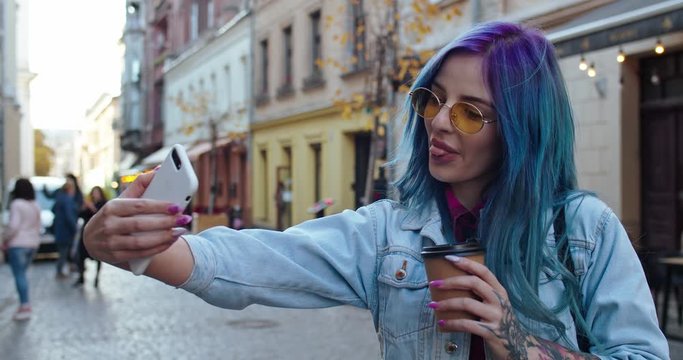 Caucasian young and cheerful girl in hipster style with blue hair posing to the smartphone camera while taking selfie photos with cup of coffee to-go at the street in the town. Close up. Outdoors.