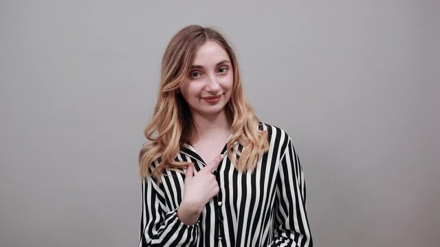Cheerful young woman in fashion black and white striped shirt pointing finger at herself, smiling isolated on gray background in studio. People sincere emotions, lifestyle concept.
