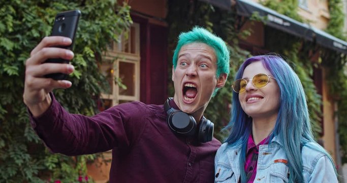 Happy and joyful young couple of Caucasian hipsters with blue hair posing funny and mocking faces to the smartphone camera while taking selfie photos outdoor.