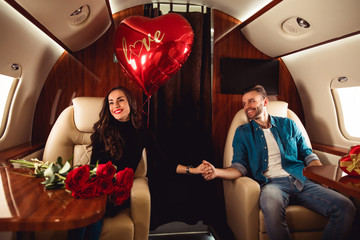 The love of our lives. A beautiful woman and an attractive man are holding hands in an aircraft cabin, celebrating St Valentine’s Day in a romantic ambiance.