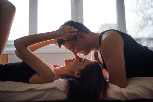 The concept of tactility, trust in a partner. A couple in love lies on the bed, legs up, laughing, hugging at home. A young man tenderly kisses a woman at the window of loft. Lovers hands touch.