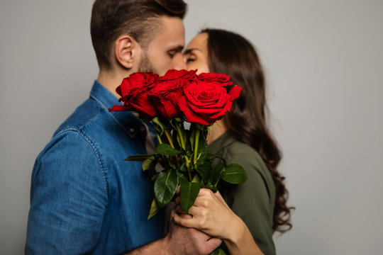 Real feelings. Close-up photo of a lovely couple in casual outfits, who are hiding behind a bouquet of red roses while kissing.