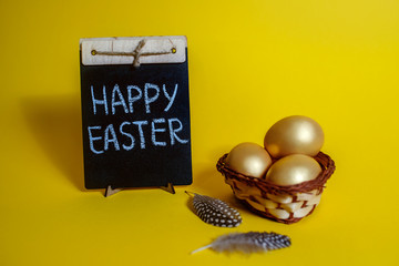 Happy Easter inscription on the blackboard, golden chicken eggs on a yellow background in a wicker basket, quail feathers decorate the Easter composition, Easter card, spring holiday