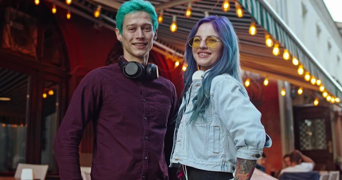 Portrait shot of the young happy Caucasian stylish couple of hipsters with blue hair standing at the stret at nice cafe terrace with garlands, hugging and smiling to the camera. Outside.
