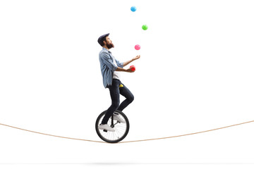Bearded male hipster juggler with balls riding a unicycle on a rope - obrazy, fototapety, plakaty