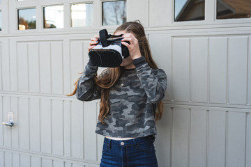 Happy/Excited teen girl holding/wearing a VR/virtual reality headset outside in front of a suburban home garage door.