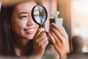 Asian smiling woman using a magnifying glass to look at the cactus in the planted pot.Concept idea of planting trees as a happy hobby.