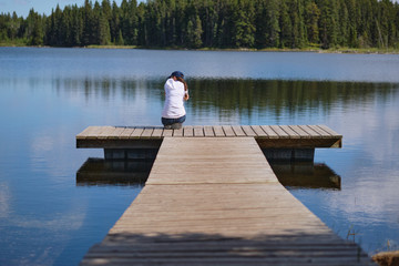 Young lady are sitting on wooden pier looking at water with forest on the background