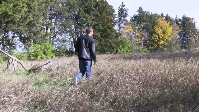Man in black leather jacket walks through long grass in the countryside.  It is a clear fall day outside of Ames, Iowa.