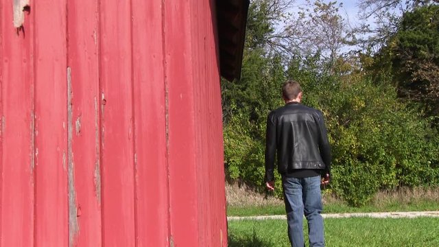 Man in black leather jacket walks besides an old red barn.  The farm is located outside of Ames, Iowa.