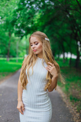 Obraz premium Portrait of a charming blond woman wearing beautiful white dress standing on the road under trees.