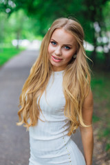 Obraz premium Portrait of a charming blond woman wearing beautiful white dress standing on the road under trees.