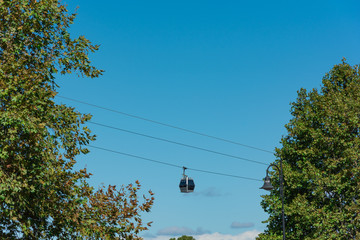 A cable railway cabin with tourists passing by between trees over background of vivid blue sky.