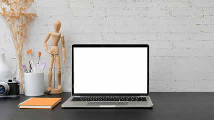 Close up view of artist workspace with blank screen laptop, wooden figure, painting brush, camera, vase and note book on black table with brick wall