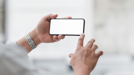 Cropped shot of  businessman touching horizontal blank screen smartphone with blurred office room
