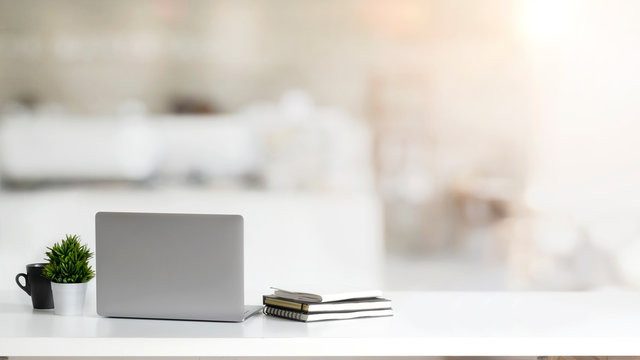 Close up view of simple workspace with laptop, notebooks, coffee cup and tree pot on white table with blurred office room