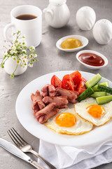 Homemade delicious breakfast with sunny side up fried egg, sausage, vegetable, jam, black coffee on a stone table. Breakfast served with flowers and white dishes