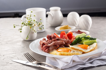 Homemade delicious breakfast with sunny side up fried egg, sausage, vegetable, jam, black coffee on a stone table. Breakfast served with flowers and white dishes