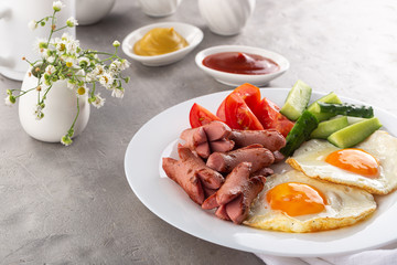 Homemade delicious breakfast with sunny side up fried egg, sausage, vegetable, jam, black coffee on a stone table. Breakfast served with flowers and white dishes