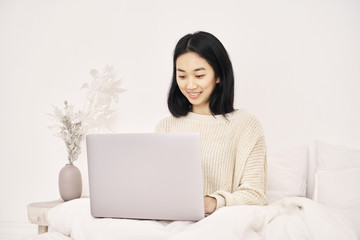 Beautiful woman freelancer Asian appearance with dark hair hairstyle Bob wears sweater has an attractive white smile works at laptop, writes post in blog about personal life career dreams of traveling
