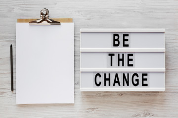'Be the change' words on a lightbox, clipboard with blank sheet of paper on a white wooden surface, top view. Overhead, from above, flat lay. Close-up.