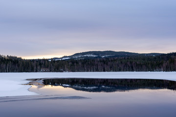 Open calm spot in a frozen lake with reflections of forest and a mountain.