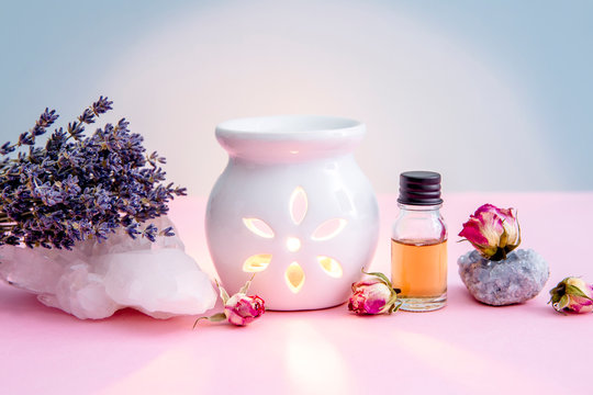White ceramic candle aroma oil lamp with essential oil bottle and dried flowers, crystal geodes on modern pastel pink and blue background indoors.