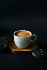 White cup of black coffee with foam on an eco wooden stand on a black background flatlay