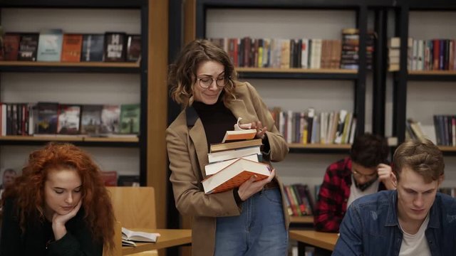 Slow motion footage of a girl in brown jacket and glasses walking by row with pile of books she took from the shelf and starting to read the book. Other students sitting, studying in college