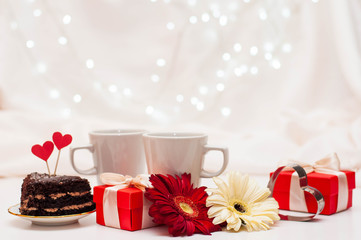 red gift boxes with ribbons, a pair of white cups and flowers on a white delicate background with bokeh.