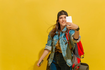 Taking selfie or vlog. Portrait of a cheerful young caucasian tourist girl with bag in jeans clothes isolated on yellow studio background. Preparing for traveling. Resort, human emotions, vacation.