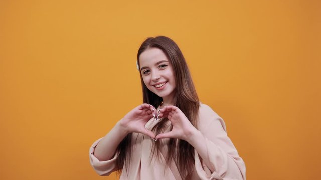 Charming caucasian brunette woman in fashion pastel shirt doing shape of heart at camera isolated on orange background in studio. People sincere emotions, lifestyle concept.