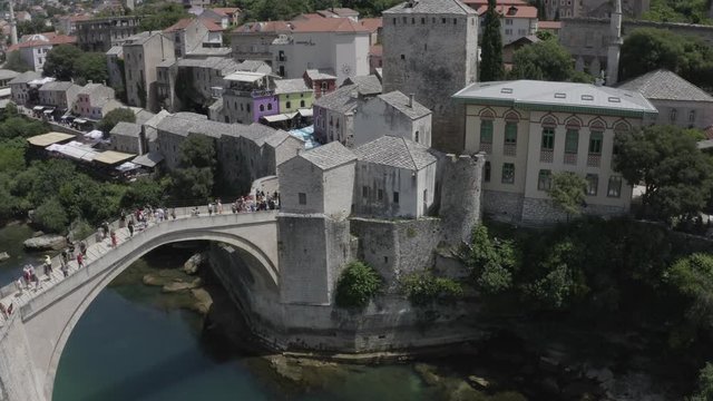 Mostar, Bosnia - June 16 2019: Aerial footage of Mostar Old Bridge (Stari Most), Bosnia and Herzegovina. Drone rises above the Neretva River and roofs of the old town. Sunny summer day.