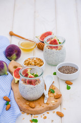 Chia pudding with yogurt, homemade granola, fresh figs and honey in a glass jar on a board on a white wooden background.