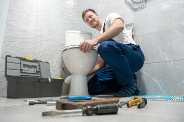 smiling man plumber in uniform repairing toilet bowl using instrument kit looks happy professional...