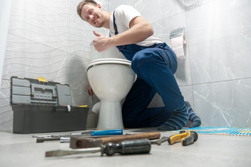 smiling man plumber in uniform repairing toilet bowl using instrument kit looks happy showing like...